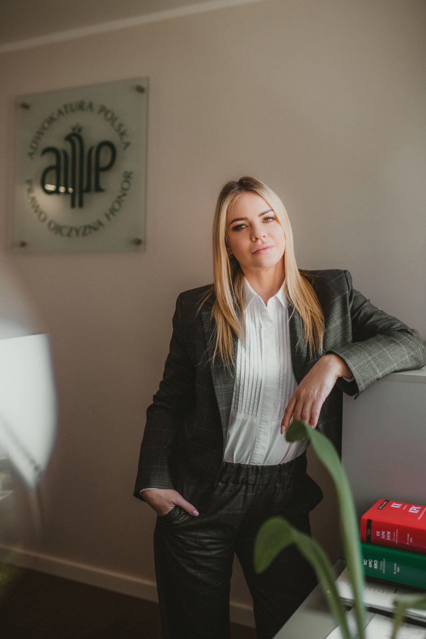 Portret kobiety w garniturze opierającej się o biurko, w tle tabliczka z logo Adwokatury Polskiej i kodeksy prawne.