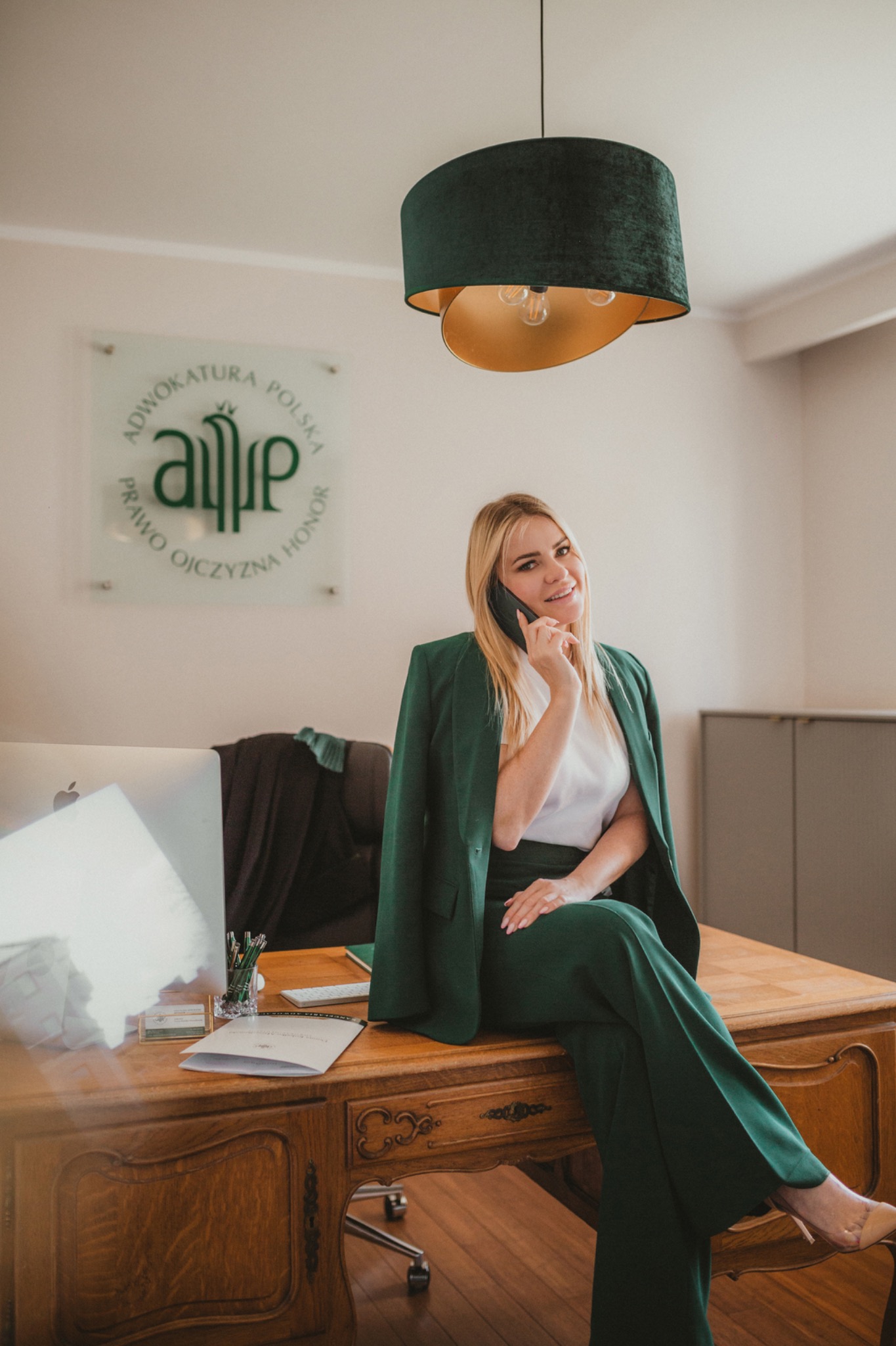 Kobieta w zielonym garniturze siedzi na biurku w gabinecie, rozmawiając przez telefon; w tle widoczne logo kancelarii i nowoczesny monitor.