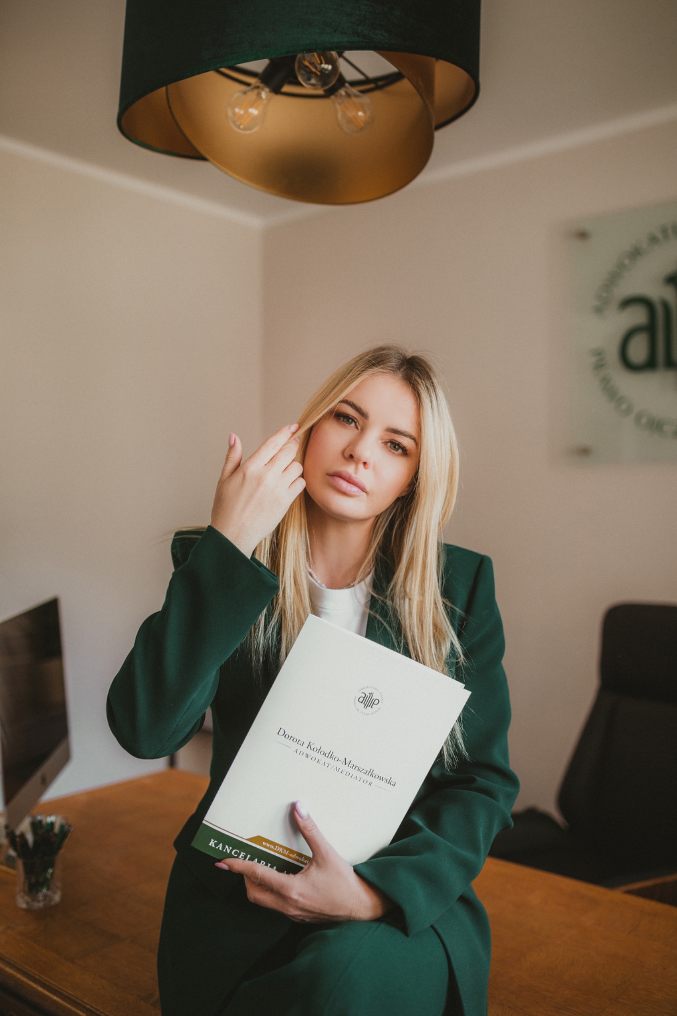 Kobieta w zielonym garniturze, siedząca przy biurku, trzymająca folder z logo kancelarii adwokackiej i danymi adwokata, w tle lampa i logo kancelarii.