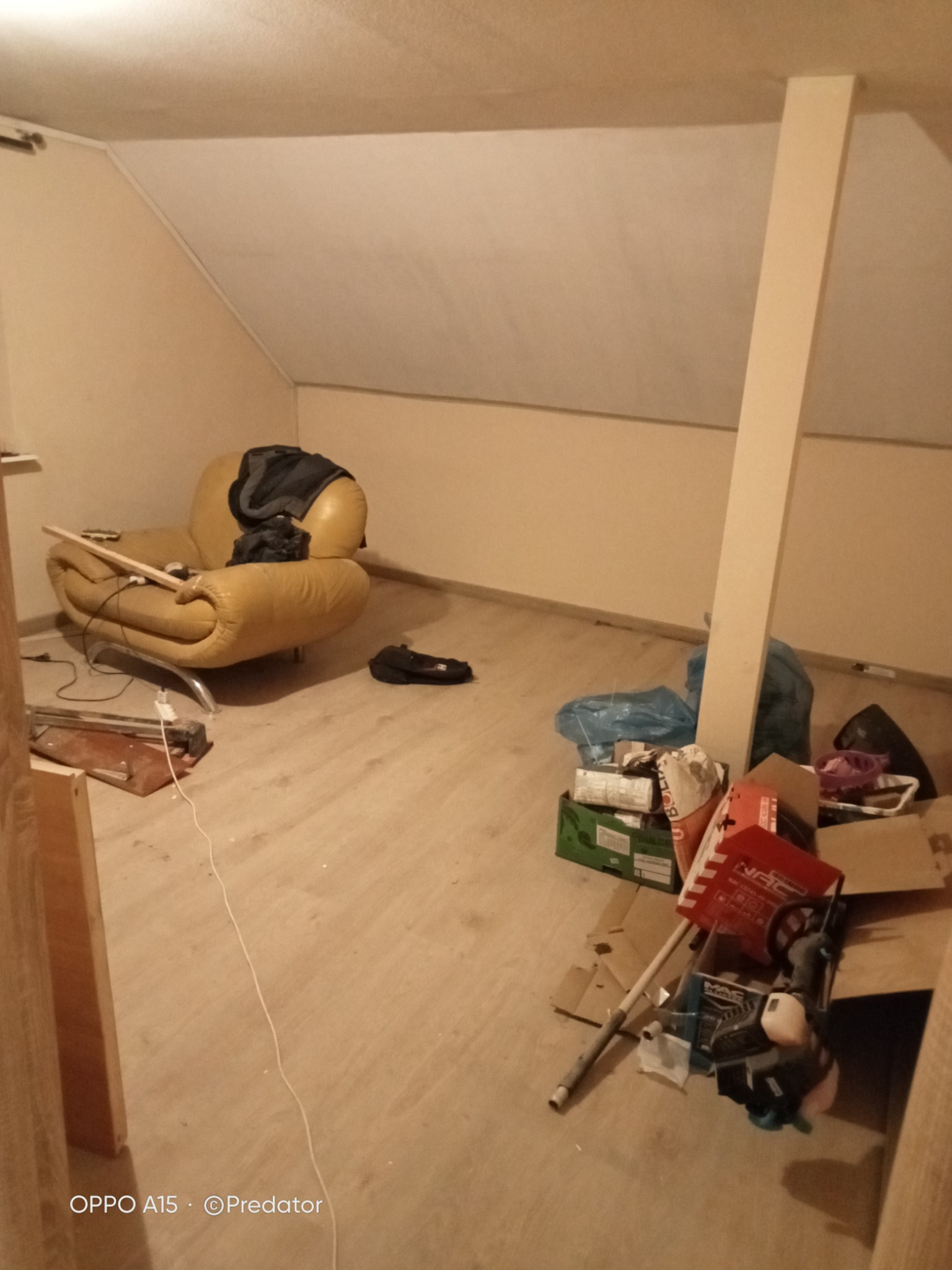 Poddasze w trakcie remontu: żółty fotel przy ścianie ze skosem, podłoga częściowo zasłonięta kartonami i workami z materiałami budowlanymi, widoczna listwa przypodłogowa oparta o ścianę.