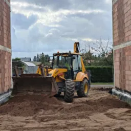 Żółta koparko-ładowarka marki Fermec na placu budowy, widoczna pomiędzy ścianami z czerwonej cegły, z wykopaną ziemią w łyżce i na ziemi, pochmurne niebo w tle.