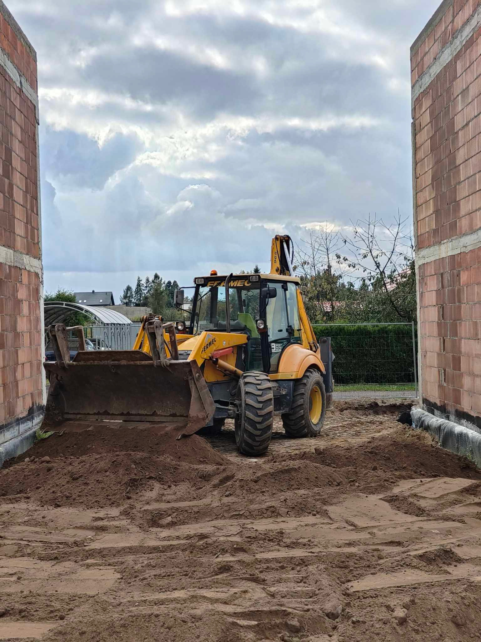 Żółta koparko-ładowarka marki Fermec na placu budowy, widoczna pomiędzy ścianami z czerwonej cegły, z wykopaną ziemią w łyżce i na ziemi, pochmurne niebo w tle.