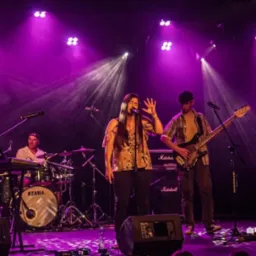 Wokalistka z długimi włosami w kwiecistej bluzce gestykuluje na scenie, obok basista w kraciastej koszuli, perkusista w tle, scena oświetlona fioletowymi reflektorami.