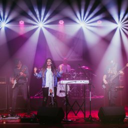 Rock Tube - Zespół muzyczny na scenie w blasku reflektorów: wokalistka, gitarzysta, basista i perkusista. Widoczne instrumenty, wzmacniacze Marshall i profesjonalne oświetlenie.