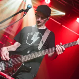 Basista w czarnej koszulce AC/DC gra na basie na scenie w czerwonym oświetleniu. Widoczny mikrofon i perkusja w tle. Muzyk ma czapkę i słuchawki.