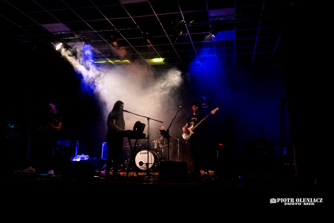 Zespół muzyczny podczas występu na scenie, oświetlony reflektorami i otoczony dymem, z widocznym perkusistą w tle.