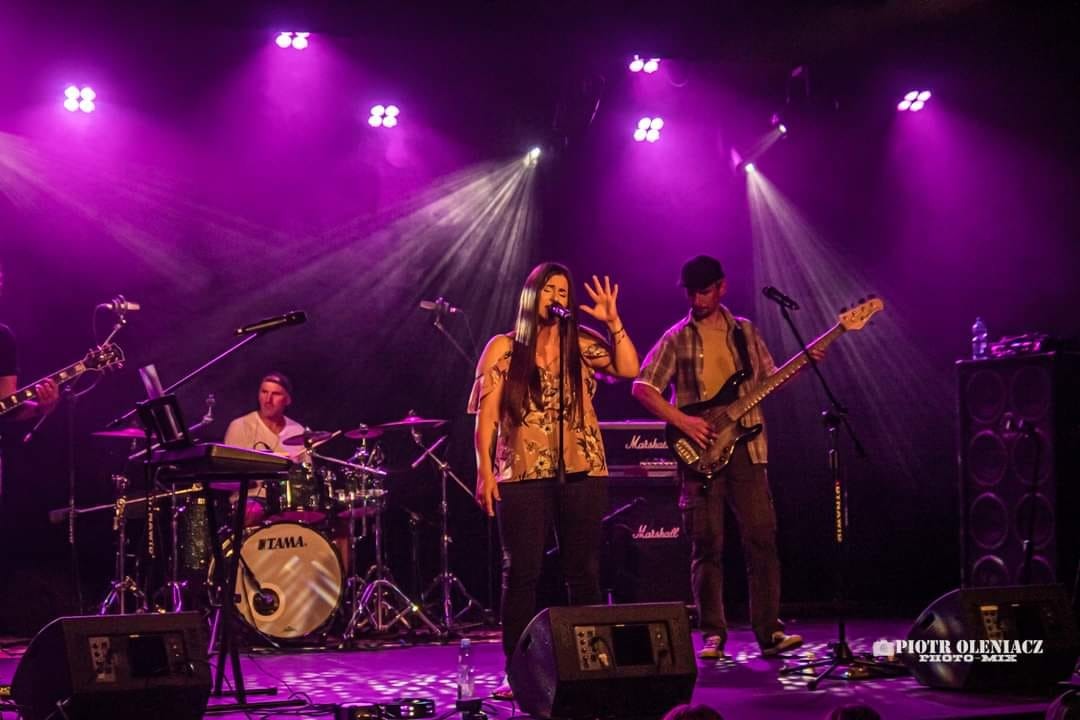 Wokalistka z długimi włosami w kwiecistej bluzce gestykuluje na scenie, obok basista w kraciastej koszuli, perkusista w tle, scena oświetlona fioletowymi reflektorami.