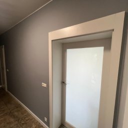 Michał Mazur - Pomieszczenie po malowaniu ścian na szaro, widoczna biała ościeżnica z zamontowanymi drzwiami z mlecznego szkła, widoczna podłoga z płytek imitujących kamień.