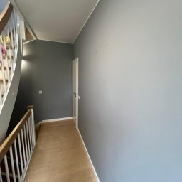 Michał Mazur - Klatka schodowa po świeżym malowaniu ścian na szaro i niebiesko, z widocznymi schodami z drewnianą barierką i panelami podłogowymi w odcieniach beżu.