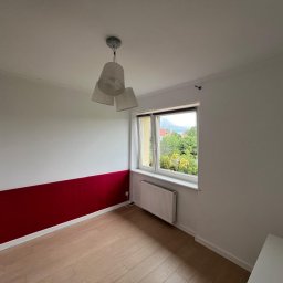 Michał Mazur - Pomalowany pokój z panelami podłogowymi, ściana podzielona na białą górę i bordowy dół, okno z widokiem na zieleń, grzejnik pod oknem, wisząca lampa z trzema abażurami.