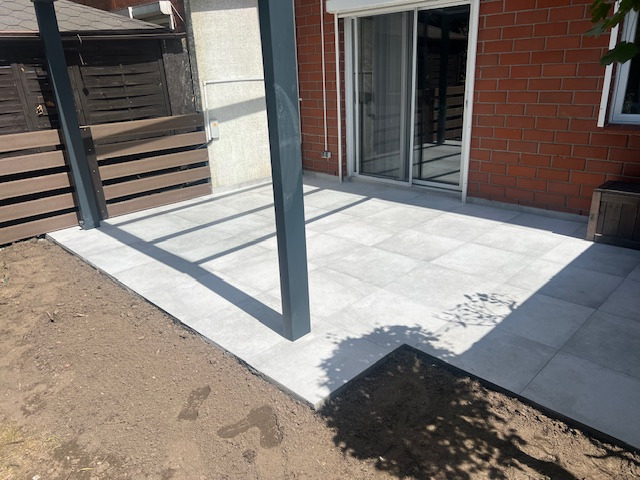 Wyłożony jasnoszarymi płytami taras z zadaszeniem, widok z poziomu gruntu, obok rabata ziemi.