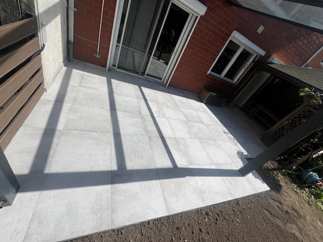 Wyłożony jasnoszarymi płytkami taras przylegający do ceglanego domu z przesuwnymi drzwiami tarasowymi i roletami, widok z góry na świeżo obsypaną ziemią rabatę.