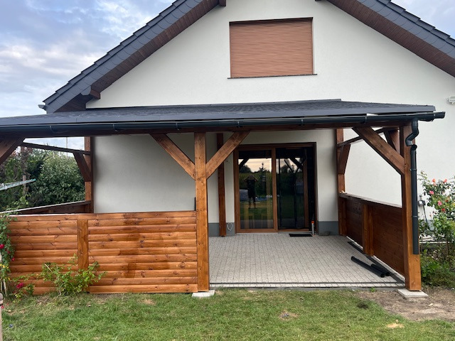 Zadaszony taras z drewnianą konstrukcją i panelami, przylegający do białego domu z brązowymi roletami, widok z zewnątrz na kostkę brukową i trawnik.