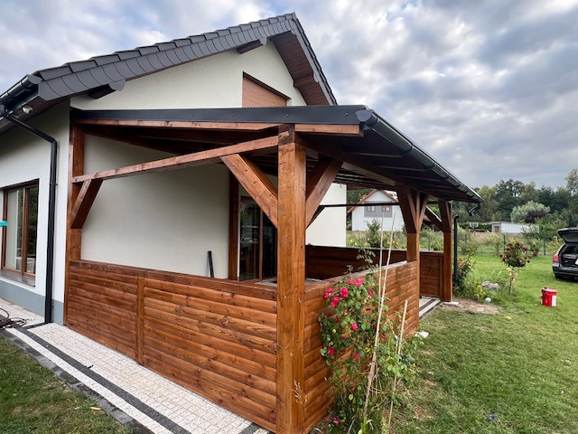 Zadaszona drewniana konstrukcja tarasu przy domu jednorodzinnym z panelami boazeryjnymi w kolorze ciemnego brązu, widok z zewnątrz, częściowo widoczny ogród z różami i samochód w tle.