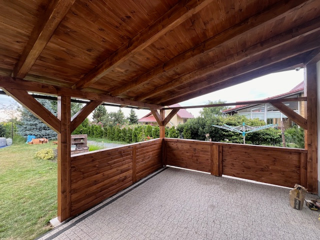 Zadaszony taras z drewnianą konstrukcją i panelami, widok na ogród z zielonym trawnikiem i drzewami, kostka brukowa na podłodze, worki z zaprawą w rogu.