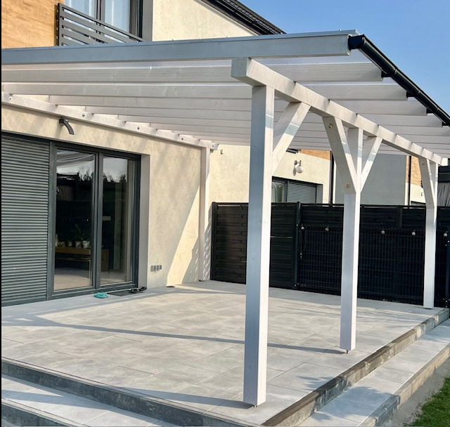 Biała drewniana pergola z zadaszeniem na tarasie z szarych płytek, widok na przesuwne drzwi tarasowe i czarne ogrodzenie.