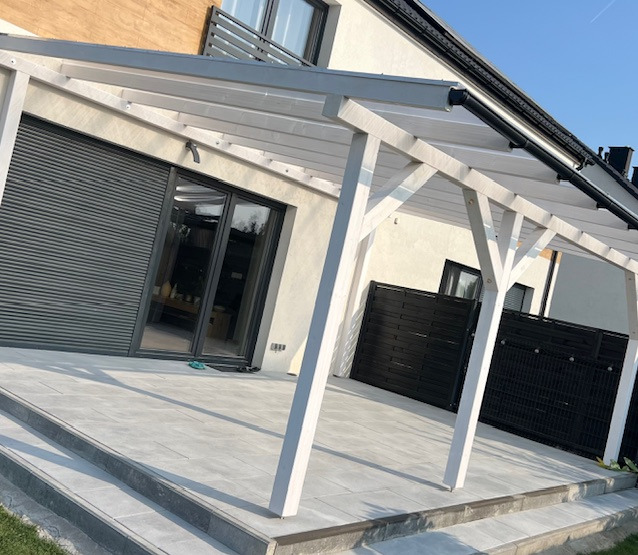 Biała, drewniana pergola z przezroczystym zadaszeniem, przylegająca do jasnego budynku z szarymi roletami w Nowym Dworze, wyłożona szarymi płytami tarasowymi.