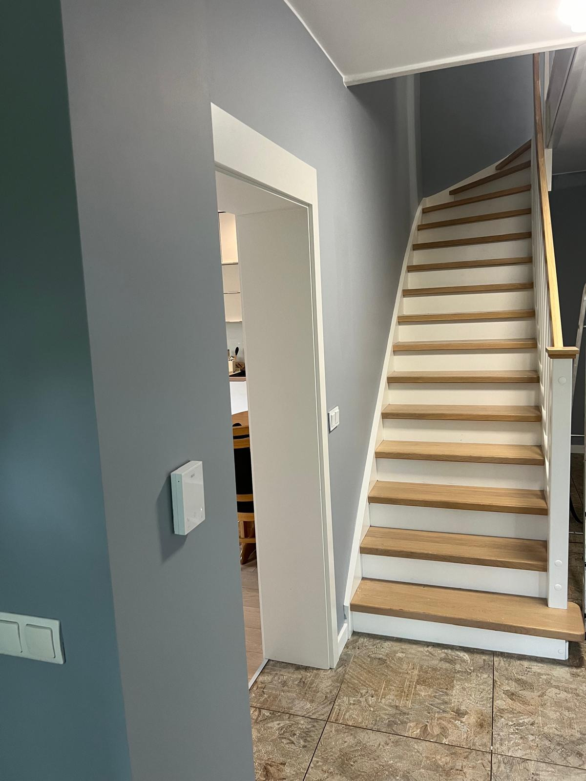 Szare wnętrze z drewnianymi schodami z białymi podstopnicami, widok na otwarte drzwi do jasnej kuchni, płytki podłogowe o wzorze marmuru.