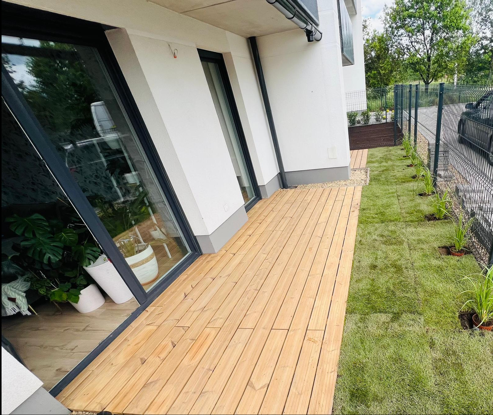 Nowoczesny taras z desek kompozytowych przylegający do budynku z dużymi oknami, obok trawnik z młodymi roślinami w doniczkach wzdłuż ogrodzenia.