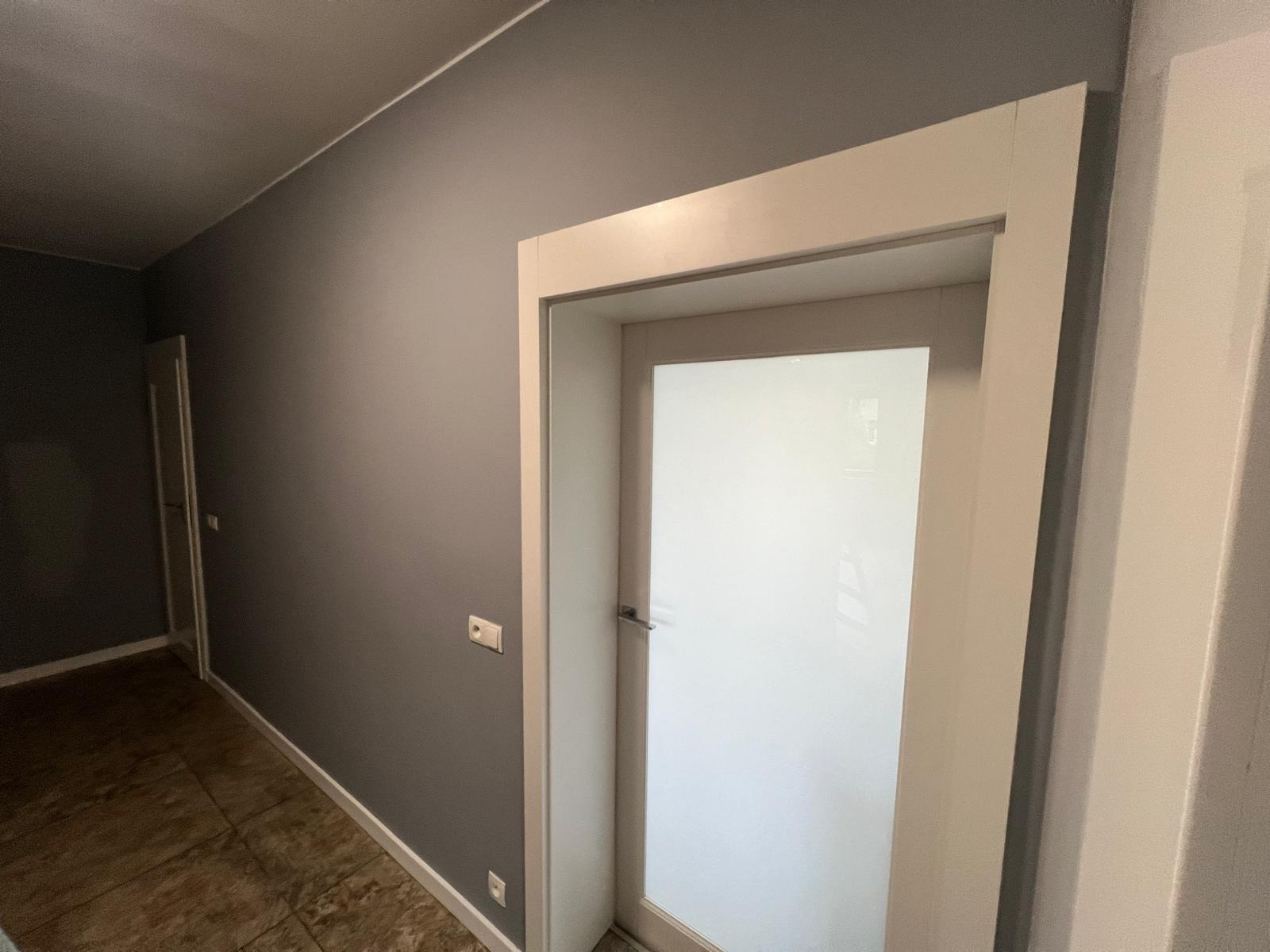 Pomieszczenie po malowaniu ścian na szaro, widoczna biała ościeżnica z zamontowanymi drzwiami z mlecznego szkła, widoczna podłoga z płytek imitujących kamień.