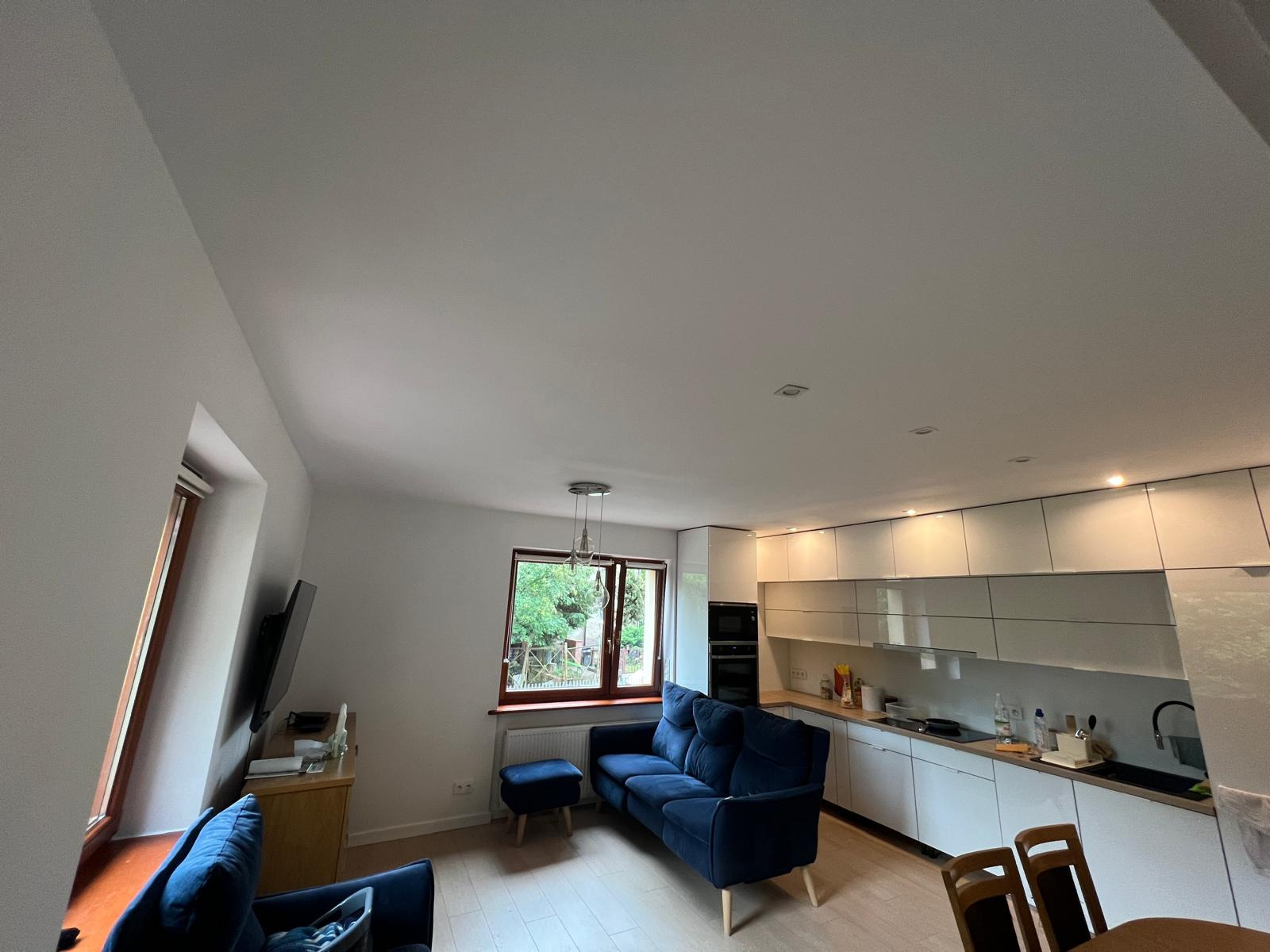 Jasne wnętrze mieszkania z aneksem kuchennym, niebieską sofą z podnóżkiem i widokiem na zieleń za oknem. Kuchnia z białymi, lakierowanymi szafkami.