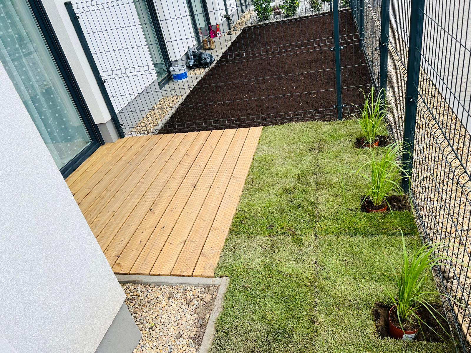 Nowoczesny taras z jasnego drewna sąsiadujący z trawnikiem i rabatą z młodymi sadzonkami, ogrodzony siatką.