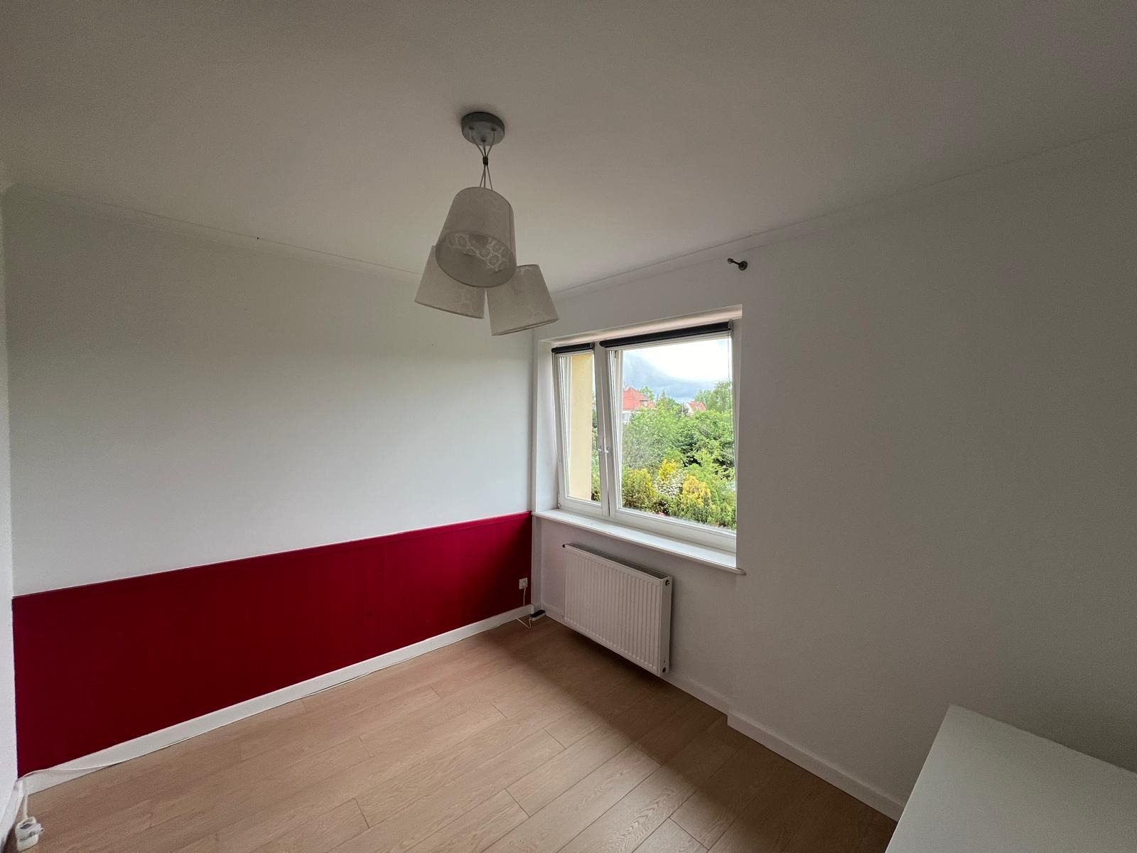 Pomalowany pokój z panelami podłogowymi, ściana podzielona na białą górę i bordowy dół, okno z widokiem na zieleń, grzejnik pod oknem, wisząca lampa z trzema abażurami.