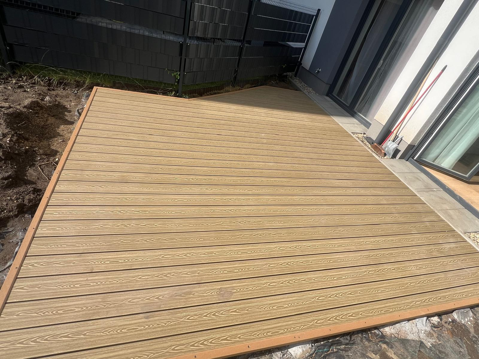 Nowo ułożony taras z desek kompozytowych o jasnobrązowym odcieniu, widok z góry, z widocznymi elementami wykończeniowymi i fragmentem ogrodu.