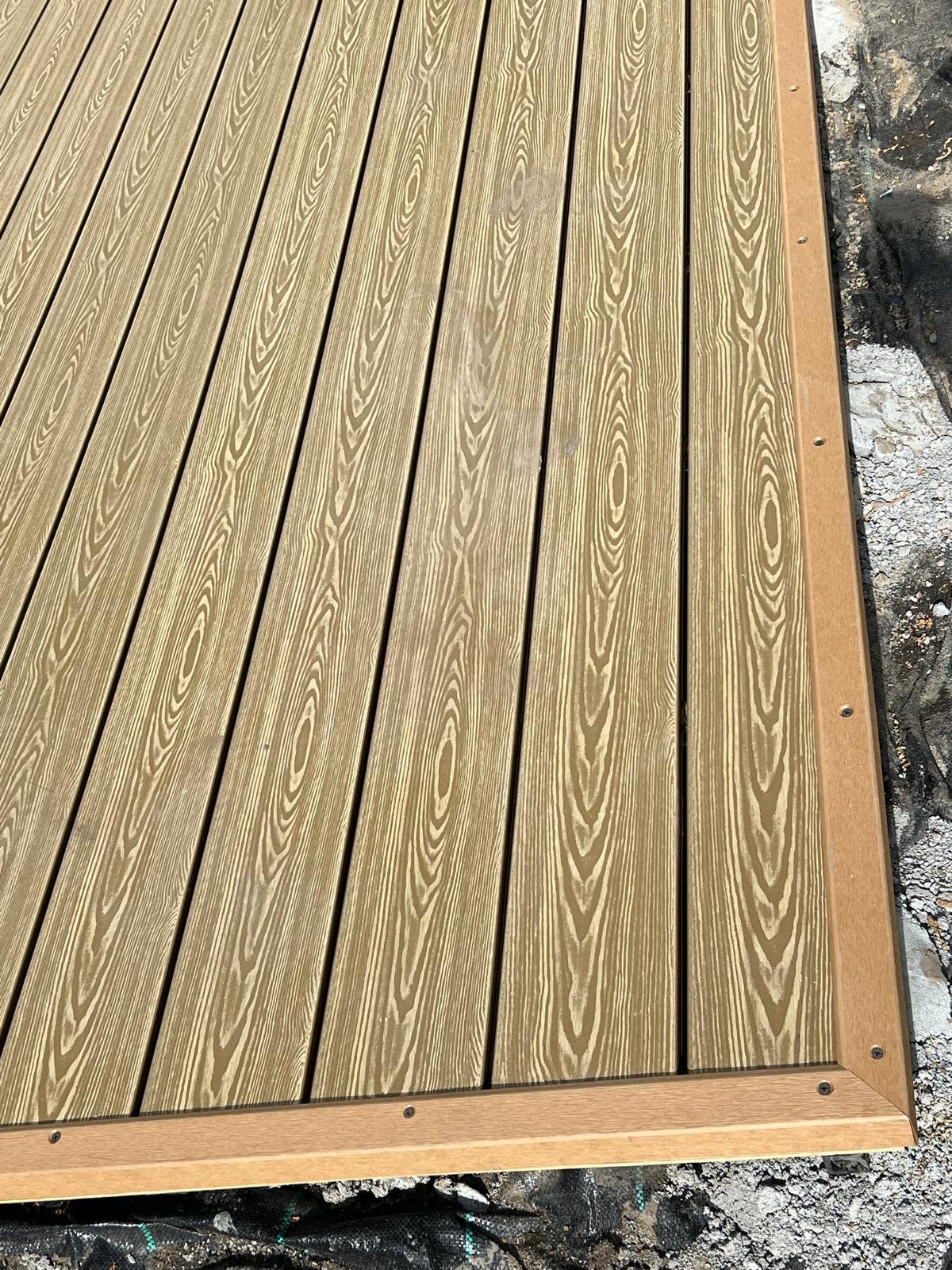 Nowo ułożona deska tarasowa kompozytowa o wyraźnym rysunku drewna, zabezpieczona brązową ramą, widoczne ślady prac ziemnych w tle.