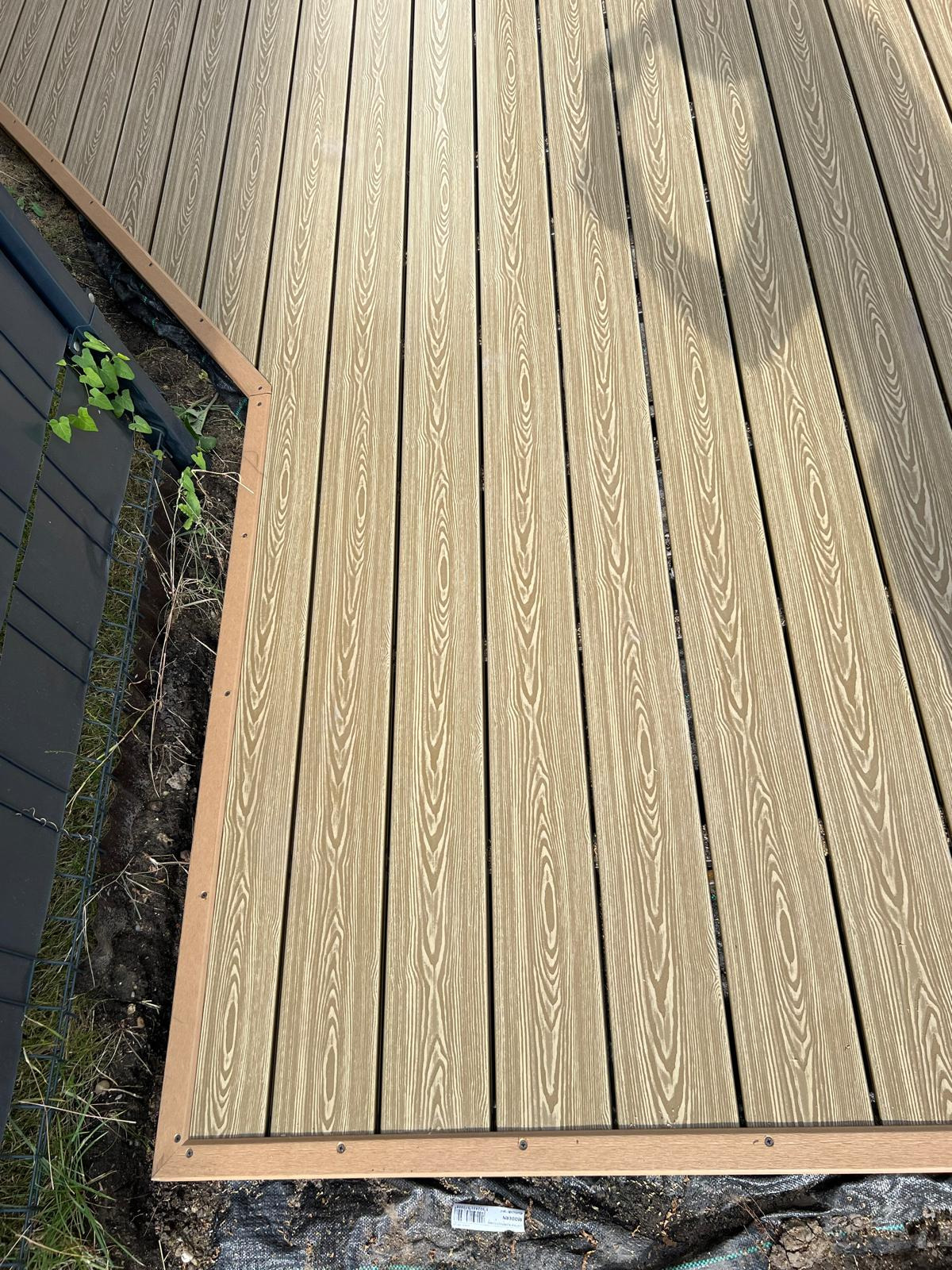 Nowo ułożony taras z desek kompozytowych w odcieniach beżu i brązu, widoczne wkręty mocujące listwy wykończeniowe, obok fragment czarnego ogrodzenia i rosnąca z ziemi zieleń.