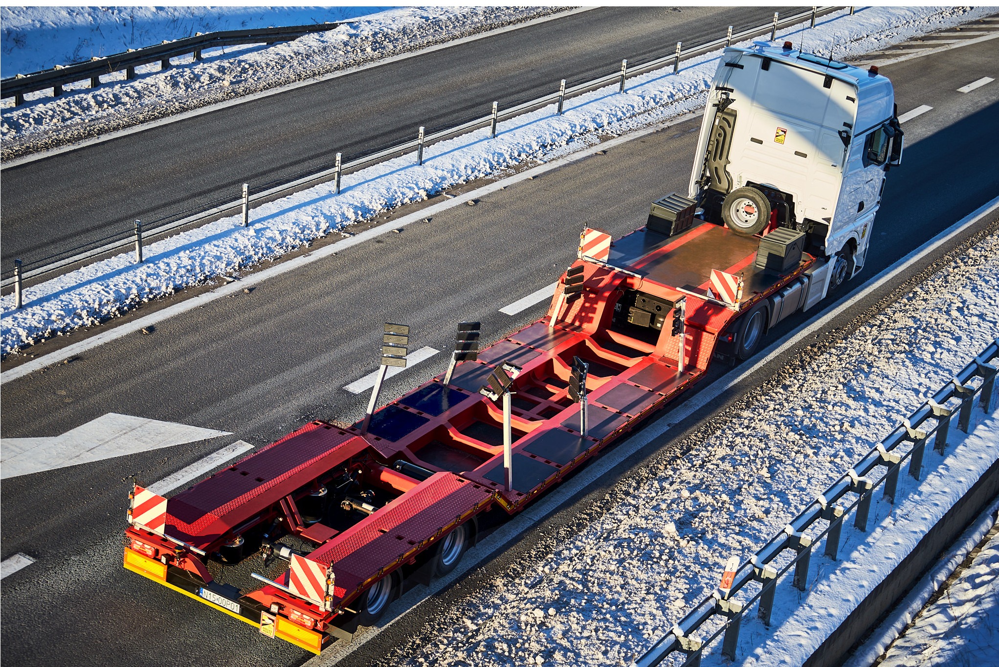 Widok z góry na czerwoną niskopodwoziową naczepę transportową z białym ciągnikiem siodłowym na zimowej autostradzie z zaspami śniegu wzdłuż barierek.