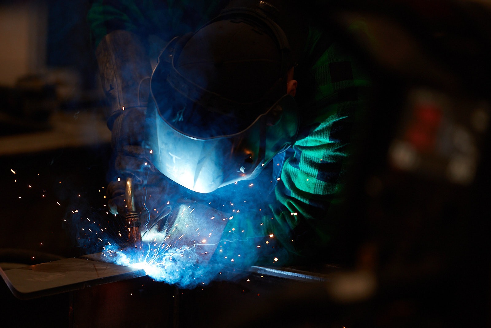 Spawacz w masce ochronnej, iskrzący łuk elektryczny podczas spawania metalu, widoczna zielono-czarna koszula w kratę.