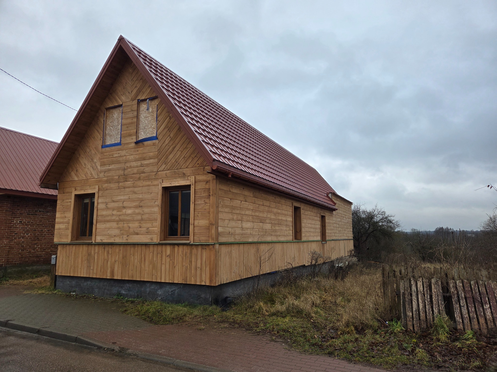 Drewniany dom z czerwonym dachem, okna zabite płytami. Widoczne deski elewacyjne i fragment chodnika. Pochmurne niebo w tle. Ujęcie z boku, z perspektywy chodnika.