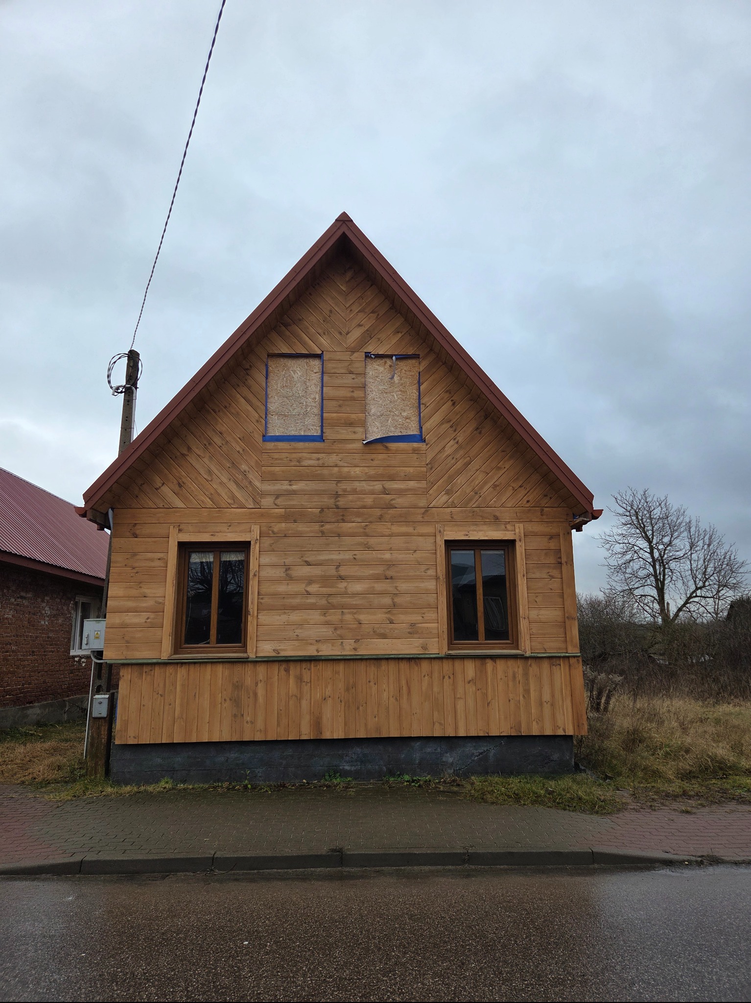 Drewniany dom z deskami w pionie, zabezpieczone okna na poddaszu, brązowy dach, widok z poziomu ulicy w pochmurny dzień.