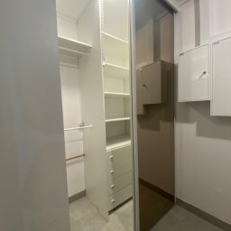 azrobieto.pl - Wnętrze garderoby z przesuwnymi drzwiami lustrzanymi, widoczne białe półki i szuflady, oświetlenie sufitowe i skrzynki instalacyjne na ścianie.