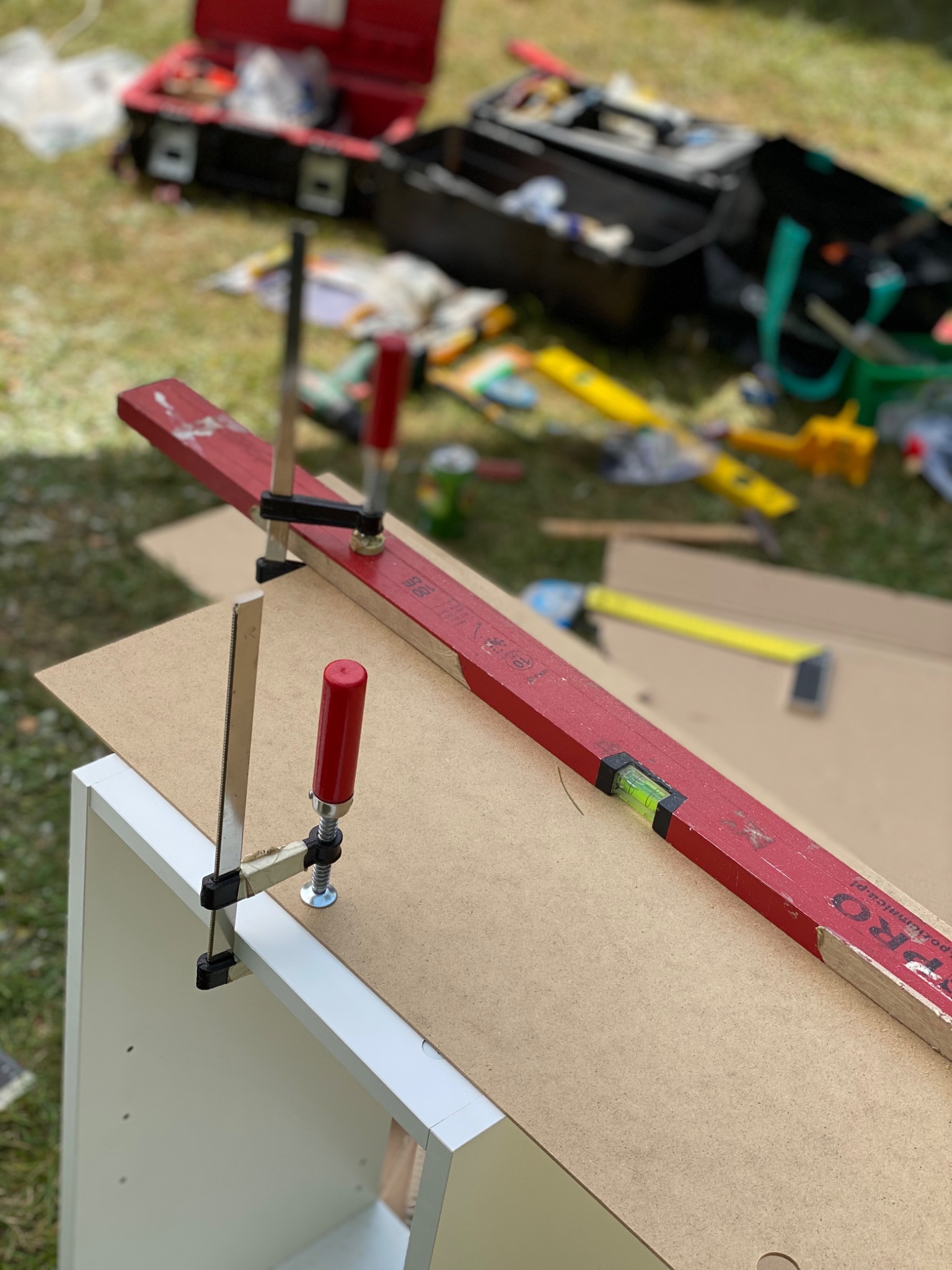 MDF płyta przymocowana do białego korpusu mebla za pomocą ścisków stolarskich i poziomicy w ogrodzie, w tle skrzynka z narzędziami.