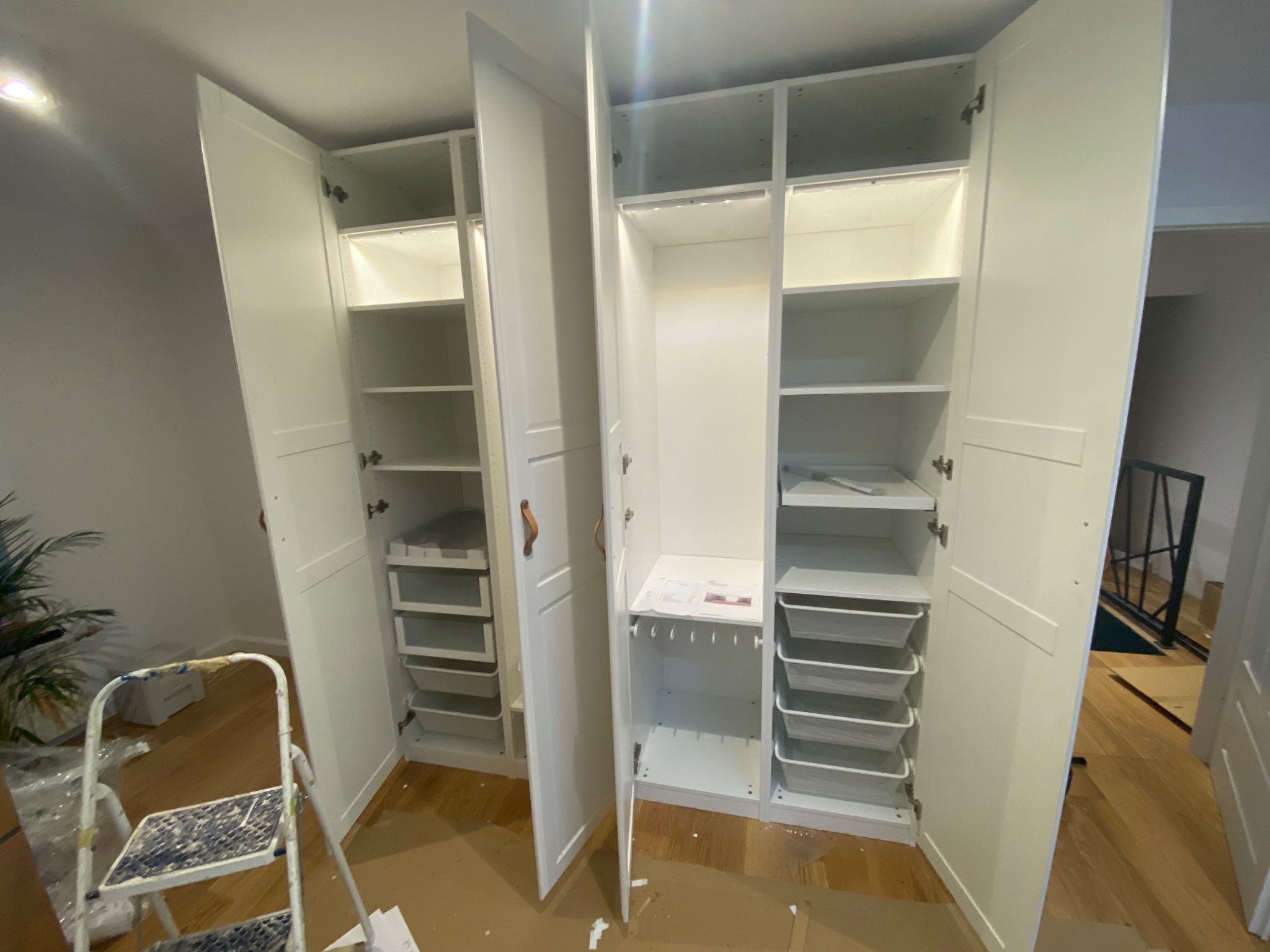 Wnętrze szafy z półkami i szufladami, oświetlone LED, widoczna drabina malarska i roślina doniczkowa w tle, podłoga zabezpieczona kartonem.