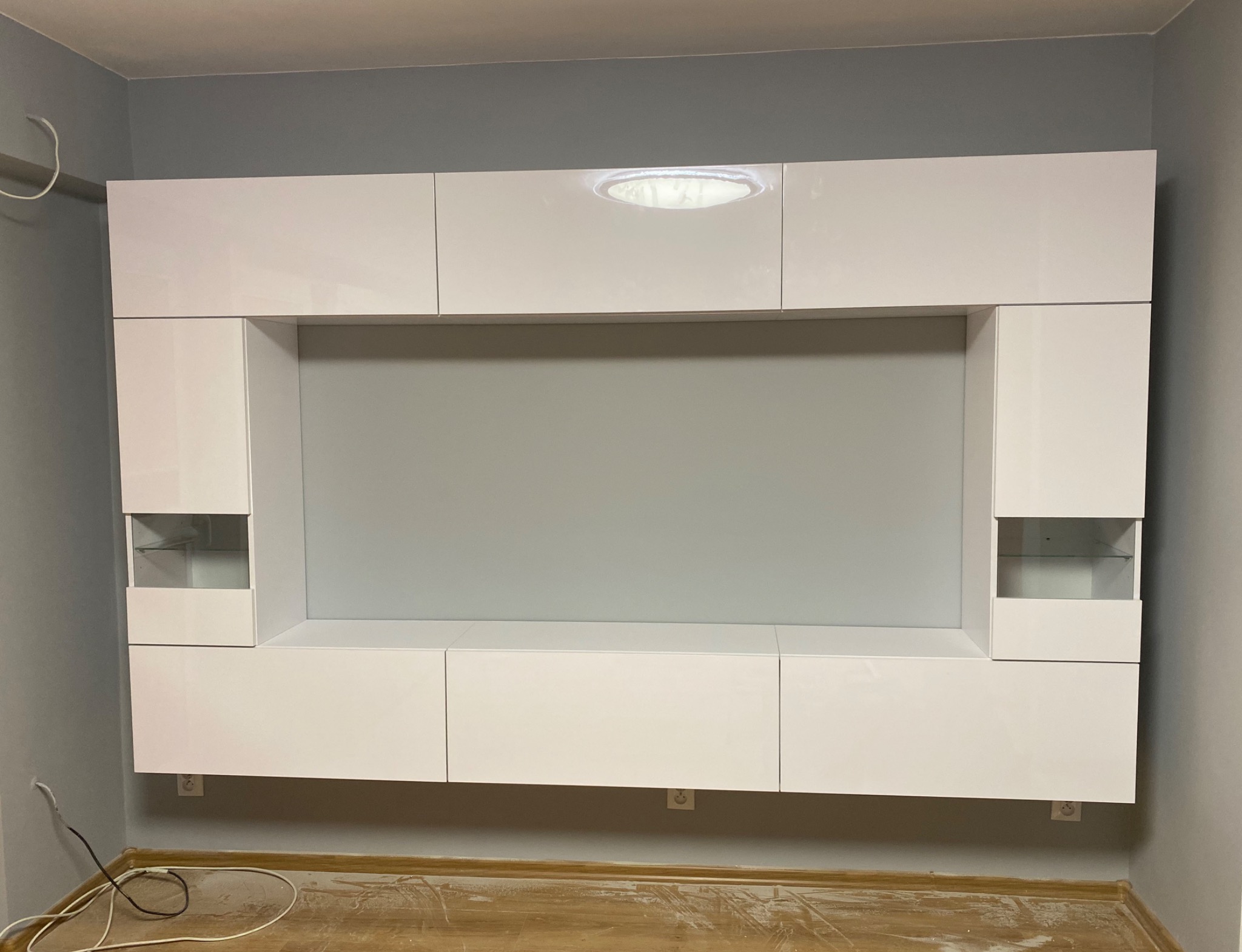 Nowoczesna, biała meblościanka z podświetleniem LED, zamontowana na szarej ścianie, widoczne gniazdka elektryczne i fragment podłogi.