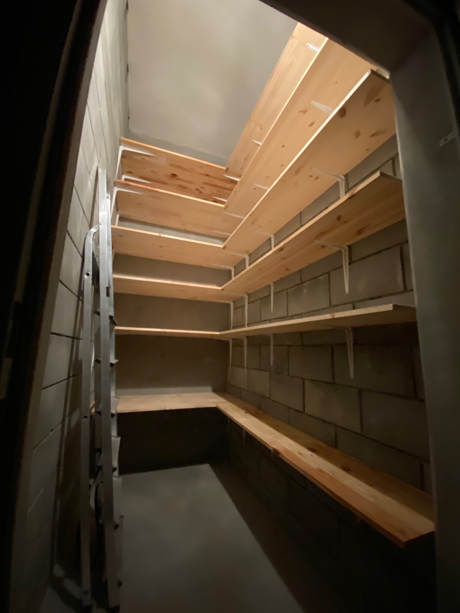 Nowo zamontowane drewniane półki na białych wspornikach w surowym pomieszczeniu z betonowych bloczków, widoczne z dołu, z drabiną opartą o ścianę.