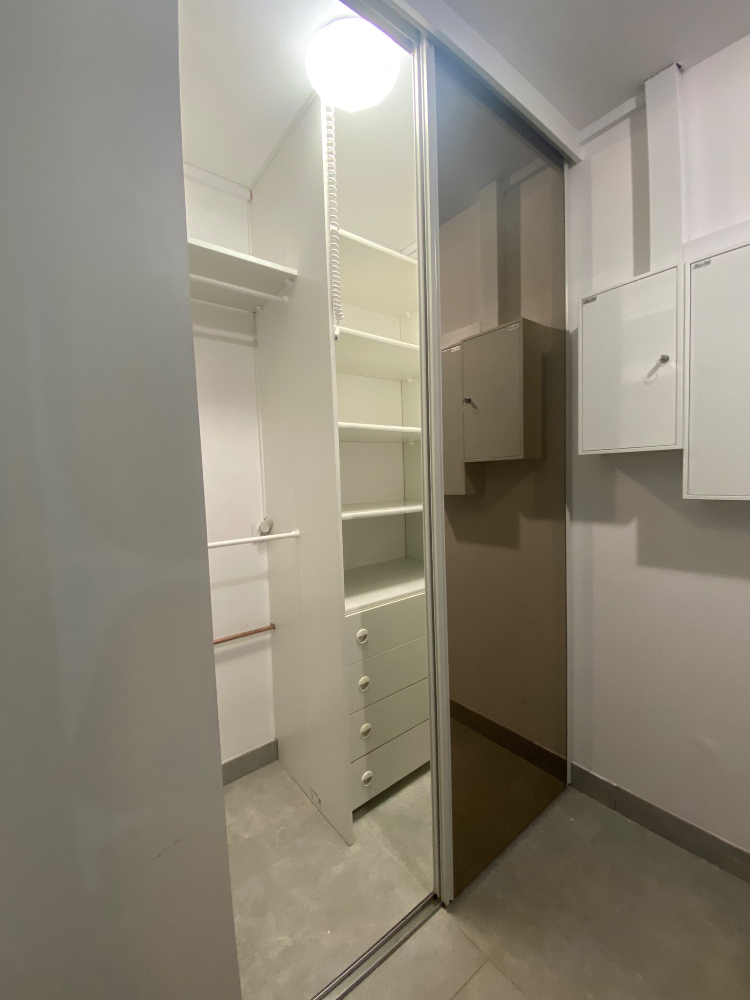 Wnętrze garderoby z przesuwnymi drzwiami lustrzanymi, widoczne białe półki i szuflady, oświetlenie sufitowe i skrzynki instalacyjne na ścianie.