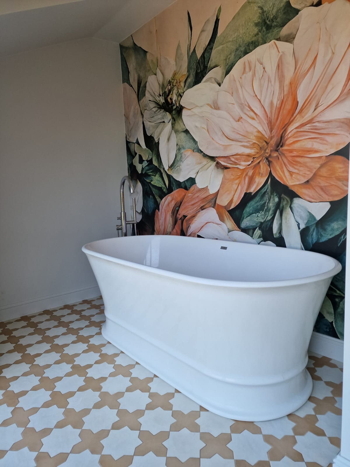 Wolnostojąca biała wanna na wzorzystej, kremowo-brązowej podłodze, z baterią podłogową i fototapetą w kwiaty w tle.