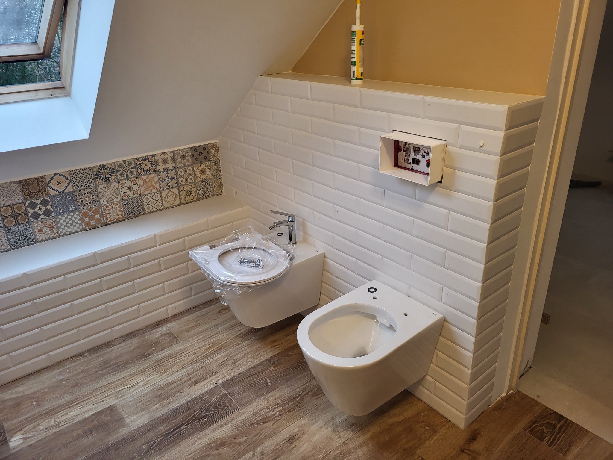 Nowa łazienka na poddaszu ze skośnym sufitem, białe kafelki typu metro, drewniana podłoga, bidet i wisząca muszla klozetowa jeszcze w folii ochronnej, widoczna puszka instalacyjna i tuba silikonu.