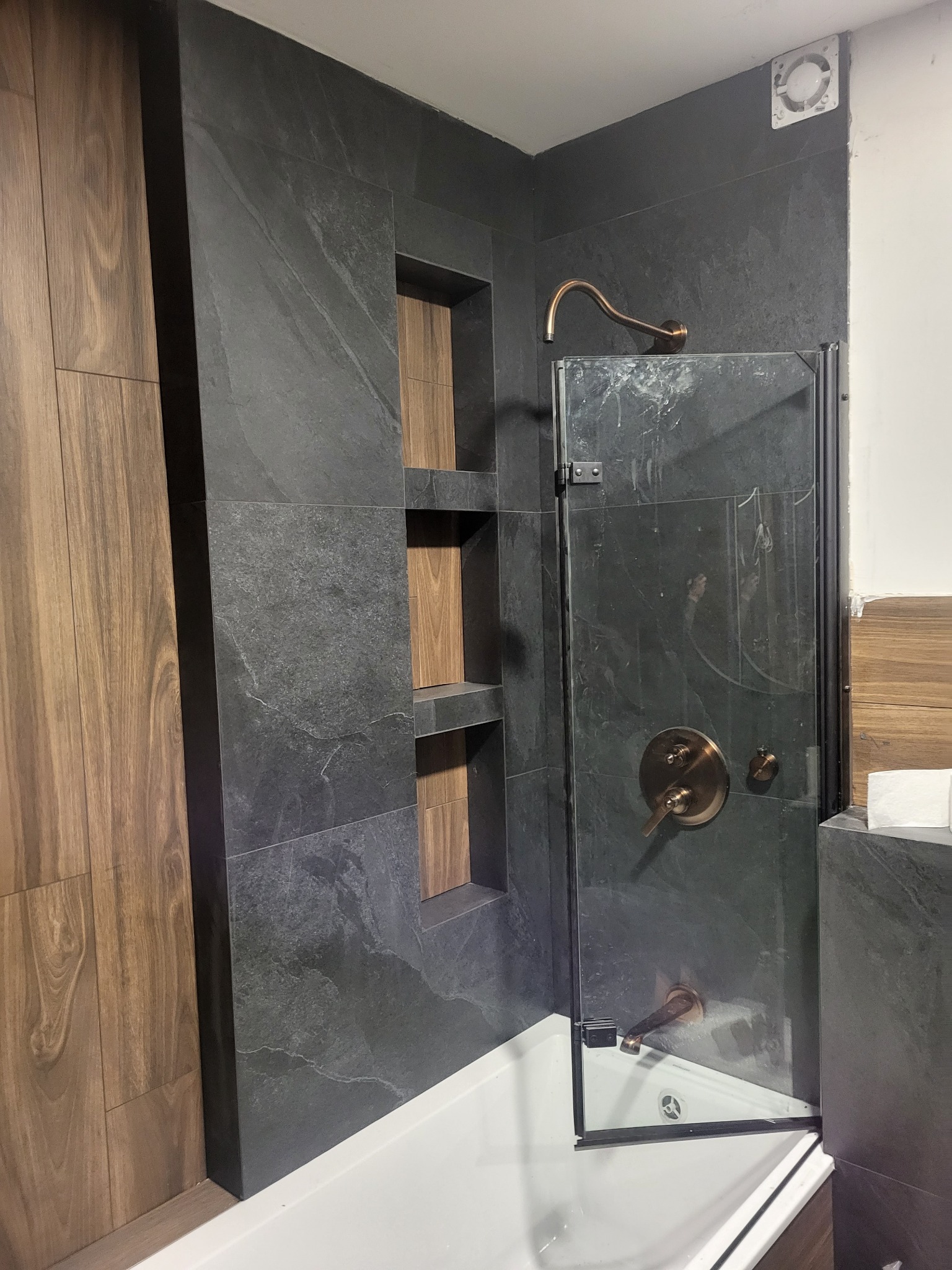 Nowoczesna łazienka z szarymi płytkami imitującymi kamień, wnękami z drewnianym wykończeniem, kabiną prysznicową ze złotą armaturą i niskim brodzikiem.