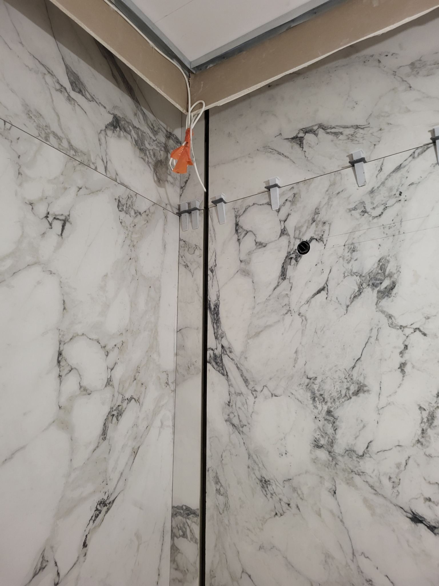 Fragment łazienki w trakcie remontu: białe płytki imitujące marmur na ścianach, system poziomowania płytek, wystające kable z pomarańczową złączką.
