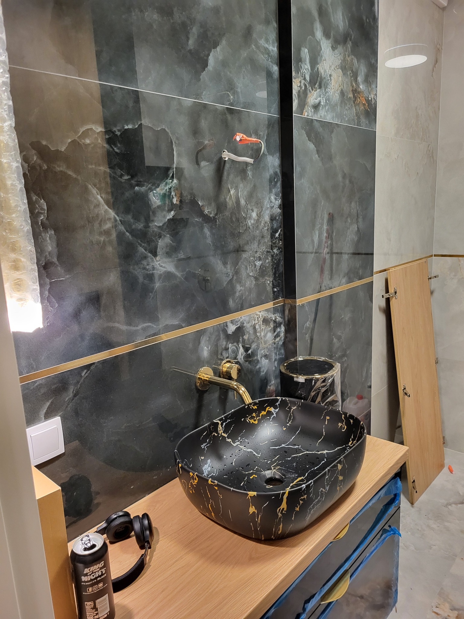 Czarna umywalka nablatowa z marmurowym wzorem i złotymi akcentami, zamontowana na drewnianym blacie, z czarnymi słuchawkami i puszką napoju obok, na tle marmurowych płytek ściennych z dekoracyjną...