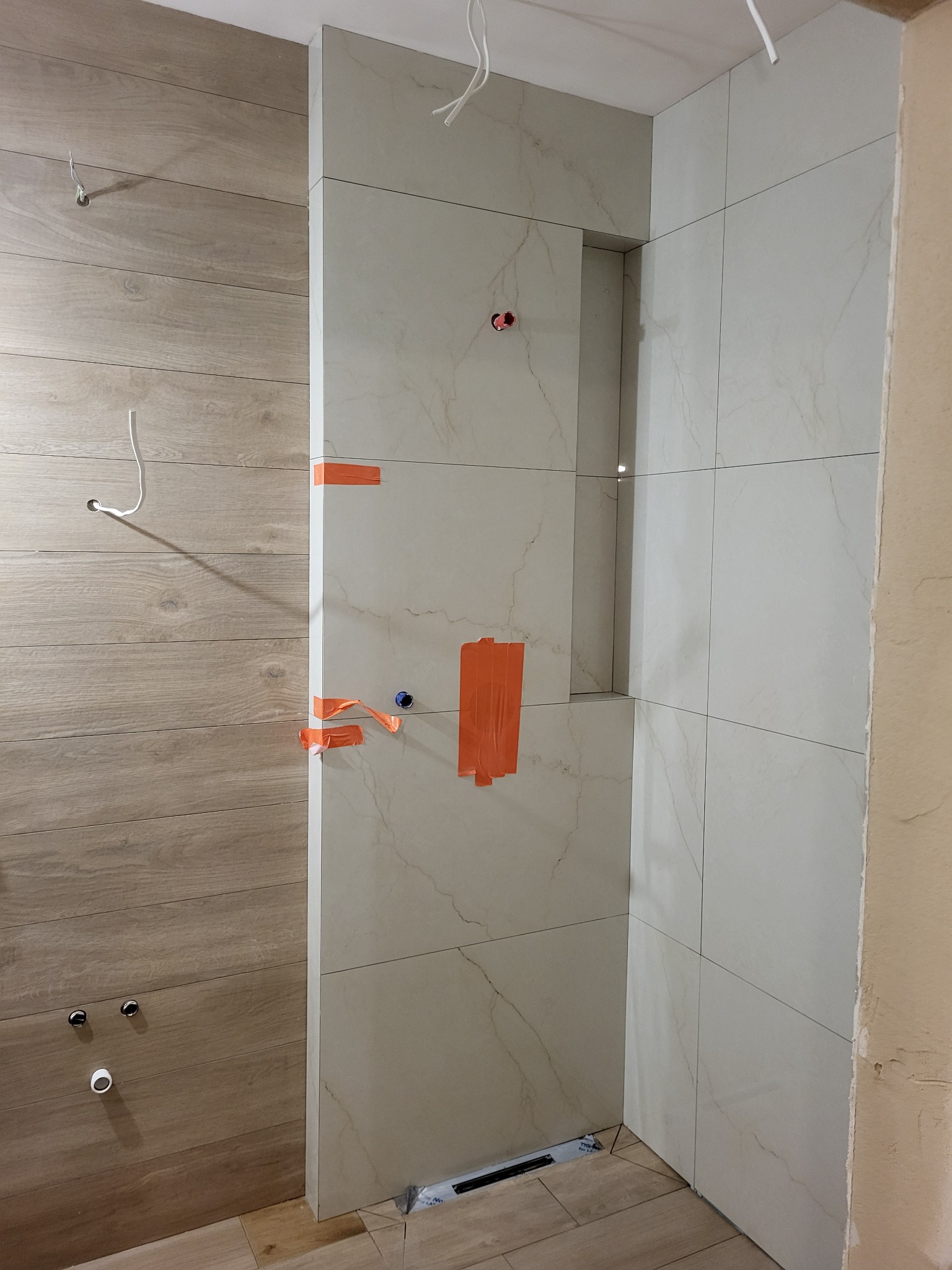 Łazienka w trakcie prac wykończeniowych; widoczne płytki imitujące drewno na ścianie i podłodze, oraz jasne płytki marmurowe na ścianach kabiny prysznicowej z wnęką, zabezpieczone taśmą malarską...