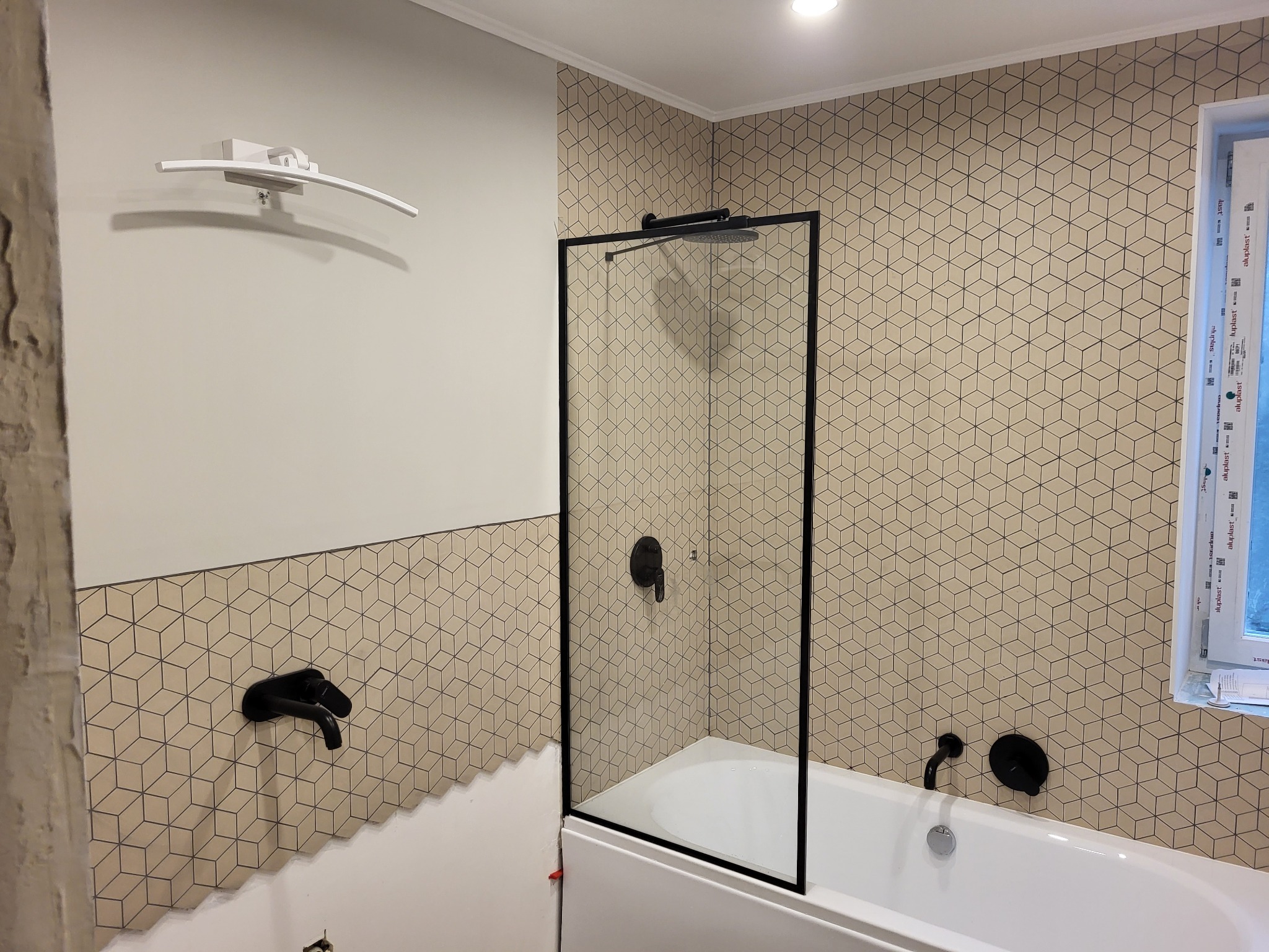 Łazienka w trakcie wykańczania: biała wanna z czarną armaturą i szklanym panelem prysznicowym na tle ściany wyłożonej beżowymi płytkami w geometryczne wzory, widoczna biała lampa nad umywalką...