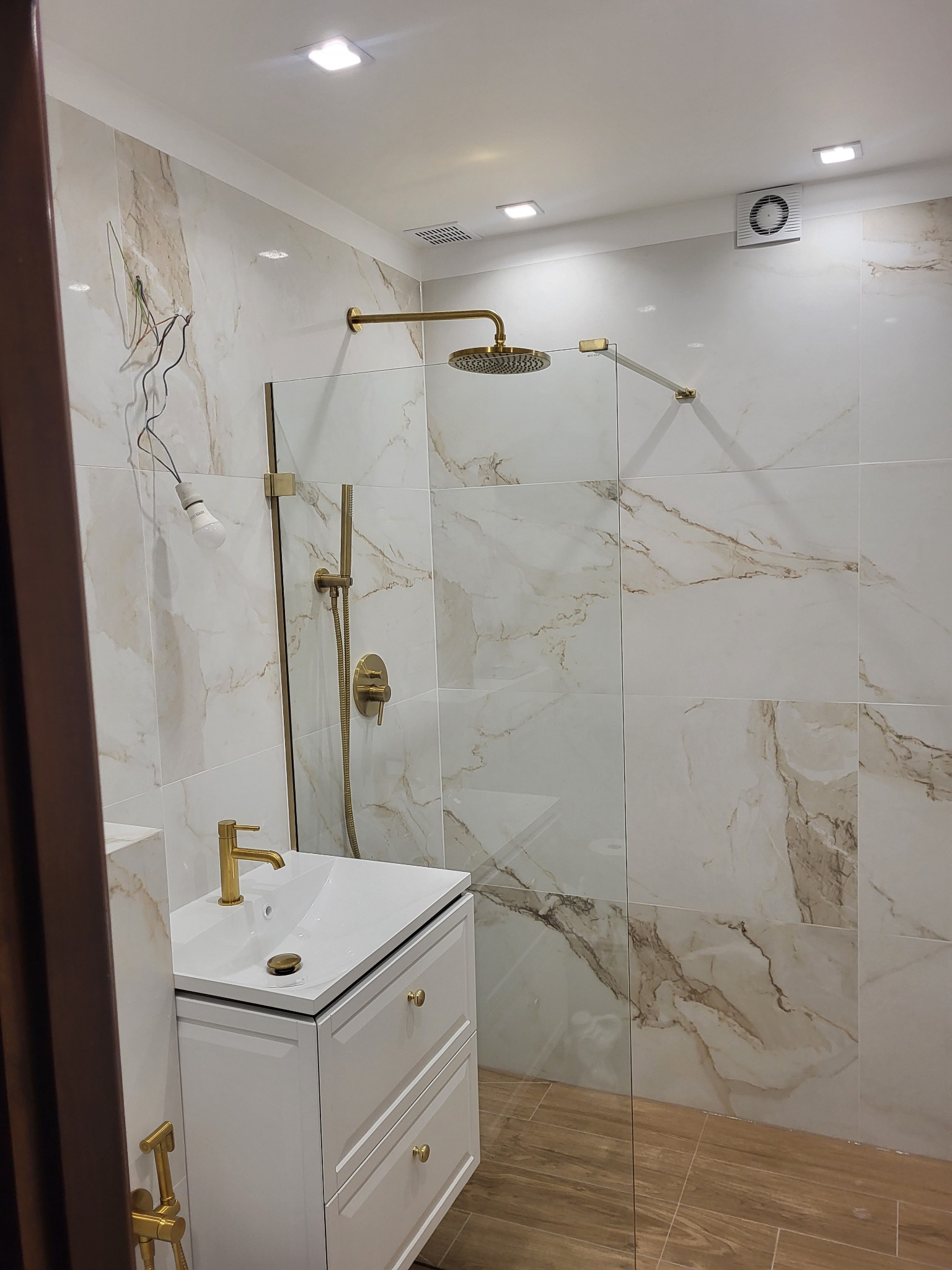 Nowoczesna łazienka w trakcie wykańczania: białe marmurowe płytki, złota armatura, kabina prysznicowa typu walk-in, biała szafka umywalkowa i niedokończone podłączenie elektryczne z żarówką.