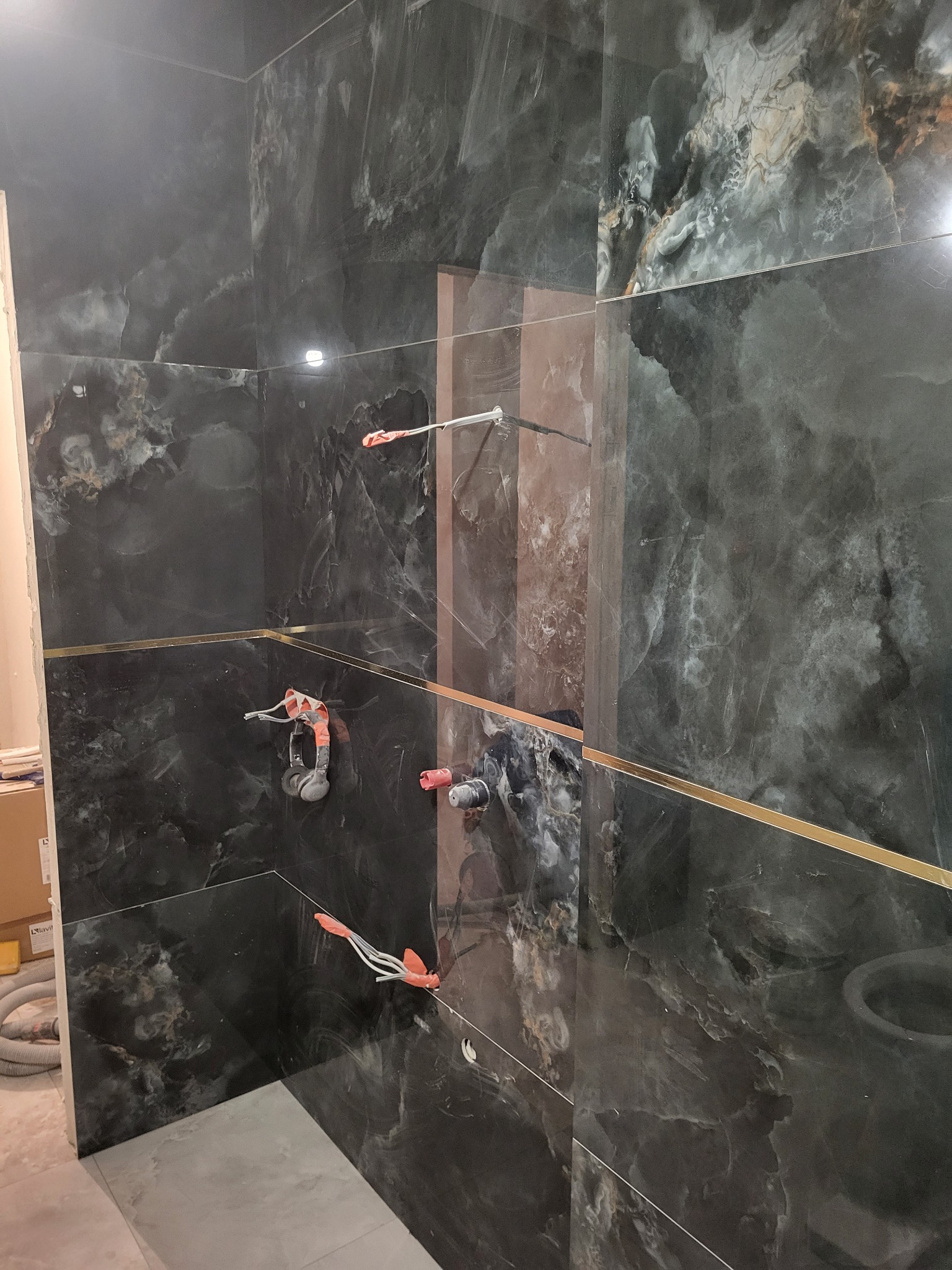 Ściana łazienki wyłożona czarnymi płytkami imitującymi marmur z delikatnymi złotymi wstawkami, widoczne wystające rury z zabezpieczeniami przed montażem armatury, szara podłoga