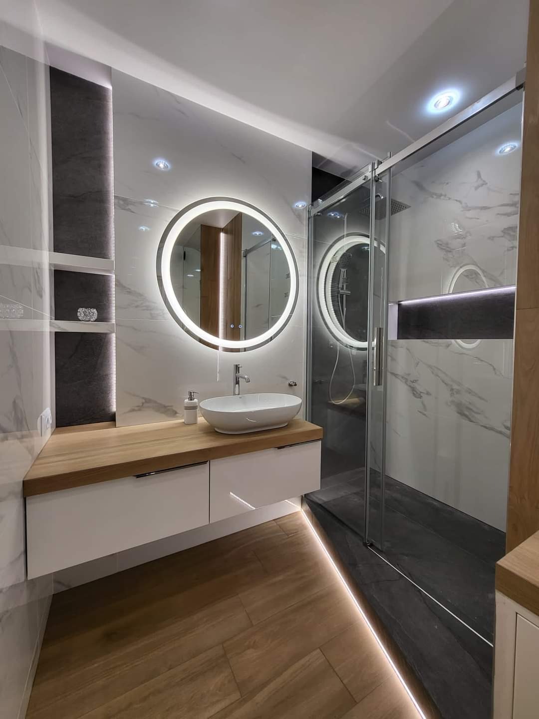 Nowoczesna łazienka z drewnianą podłogą, okrągłym lustrem z podświetleniem LED, białą umywalką nablatową i kabiną prysznicową typu walk-in z marmurowymi płytkami.